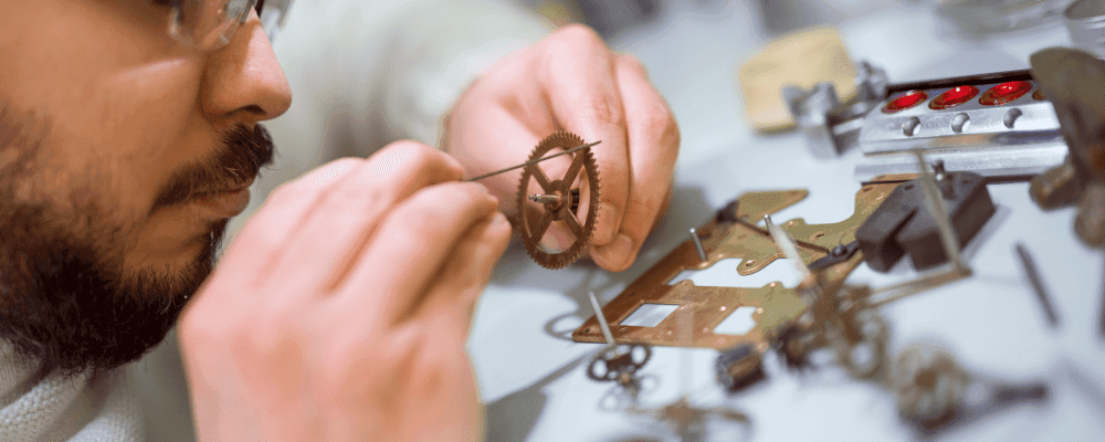 Auf diesem Bild ist eine Person zu sehen, die eine Ausbildung zum/r Uhrmacher/in absolviert. Sie sitzt an einem Tisch mit vielen kleinen Teilen und Instrumenten um sich herum. Die Person sieht konzentriert und geschickt aus, wie sie an den winzigen Teilen arbeitet.