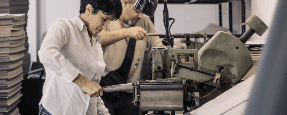 Auf diesem Bild ist eine Person zu sehen, die eine Ausbildung zum/r Buchbinder/in absolviert. Dabei handelt es sich um einen kreativen und anspruchsvollen Job, bei dem man stets mit neuen Ideen und Konzepten arbeitet. Die Buchbinder/in ist dafür verantwortlich, Bücher sowie andere Druckerzeugnisse herzustellen und zu binden. Dies erfordert viel Fingerspitzengefühl, Konzentration und Geduld.