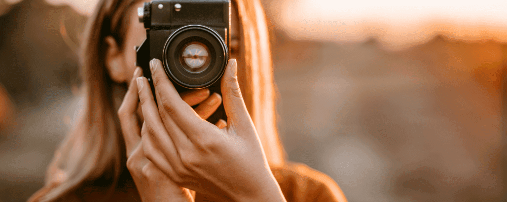 Auf diesem Bild ist eine Person zu sehen, die eine Ausbildung zum/r Fotografen/in absolviert. Die Person hält eine Kamera in der Hand und sieht damit auf das Motiv, welches sie fotografieren möchte. 