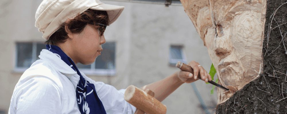 Das Bild zeigt eine Person, die eine Ausbildung zum/r Holzbildhauer/in absolviert. Die Person ist mit allen notwendigen Werkzeugen und Materialien ausgestattet und bereit, ihr Handwerk auszuüben. Die Umgebung ist ruhig und idyllisch, perfekt für die konzentrierte Arbeit am Holzblock.