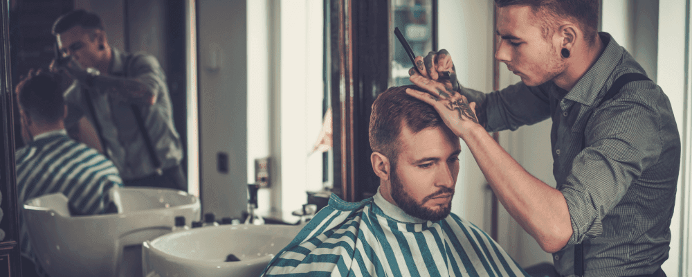 Das Bild zeigt eine Person, die eine Ausbildung zum/r Friseur/in absolviert. Die Person ist konzentriert bei der Arbeit und sorgfältig dabei, einem Kunden die Haare zu schneiden.